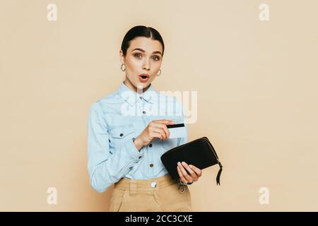 donna bruna scioccata in camicia in denim con portafoglio e credito scheda isolata su beige Foto Stock