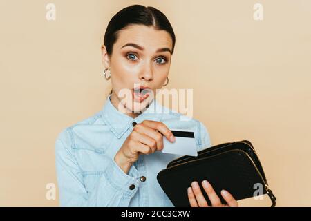 donna bruna scioccata in camicia in denim con portafoglio e credito scheda isolata su beige Foto Stock