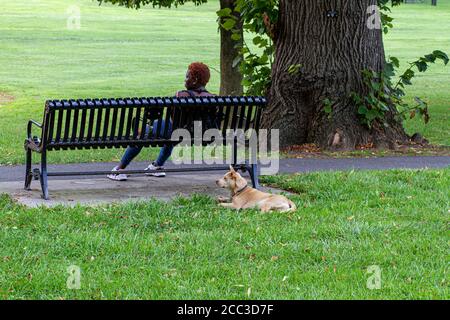 Frederick, MD, USA 08/14/2020: Una giovane donna afroamericana con capelli corti rossi che indossa tights e sneakers è seduta da sola su una panchina a Ba Foto Stock