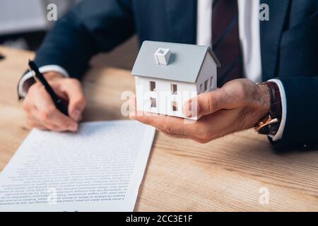 vista ritagliata di avvocato che tiene il modello di casa mentre firma i documenti sul tavolo Foto Stock