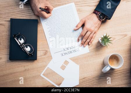 vista dall'alto dell'avvocato che firma l'accordo di polizza di assicurazione vicino a pianta, occhiali, notebook e modello della casa della tazza di carta Foto Stock