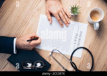 vista ritagliata di avvocato firma polizza di assicurazione accordo vicino a pianta, occhiali, notebook e stetoscopio sulla scrivania Foto Stock