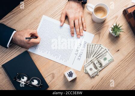 vista dall'alto dell'avvocato che firma l'accordo di polizza assicurativa vicino a pianta, occhiali, notebook, dollari e cubo con casa Foto Stock