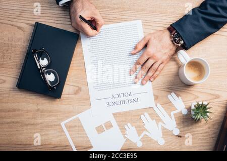 vista dall'alto dell'avvocato che firma il contratto di assicurazione vicino alla pianta, gli occhiali, il notebook e la tazza di carta modello della casa con la famiglia Foto Stock