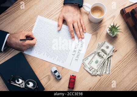 vista dall'alto dell'avvocato che firma l'accordo di polizza assicurativa vicino a pianta, occhiali, taccuino, dollari e macchinine giocattolo Foto Stock