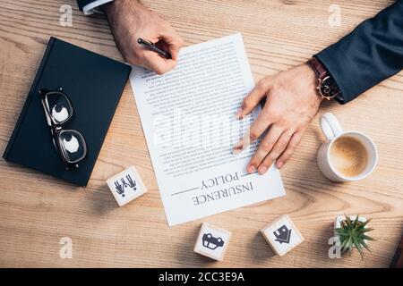 vista dall'alto dell'avvocato che firma l'accordo di polizza assicurativa vicino a pianta, occhiali, taccuino e cubetti di legno con la famiglia, l'automobile e la casa Foto Stock