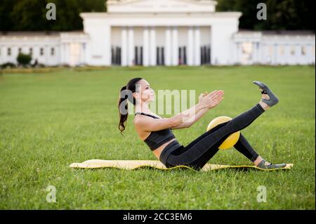 Bella giovane donna sdraiata su un materasso giallo, posa mentre indossa un vestito sportivo stretto nel parco facendo pilates o yoga, teaser con palla exercis Foto Stock