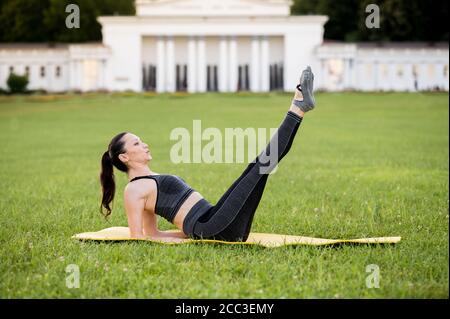 Bella giovane donna sdraiata su un materasso giallo, posa mentre indossa un vestito sportivo stretto nel parco facendo pilates o yoga, esercizi di torsione dell'anca Foto Stock