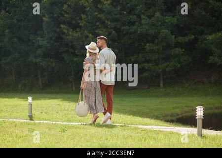 Vista posteriore ritratto di una romantica coppia adulta che si abbraccia mentre si cammina sul sentiero attraverso il prato verde in uno scenario naturale, copia spazio Foto Stock