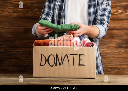 vista a taglio dell'uomo che mette i vestiti in scatola di cartone con scritta donate su sfondo di legno, concetto di carità Foto Stock