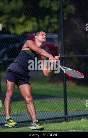 MD, USA 08/14/2020: Un atletico ispanico che indossa una camicia e un pantaloncini senza maniche sudati sta prendendo un servizio durante una partita di tennis in un campo locale a Ba Foto Stock