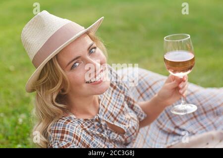 Ritratto di bella donna bionda sorridente alla macchina fotografica e sdraiata su erba verde mentre si gode pic-nic all'aperto, copy spazio Foto Stock