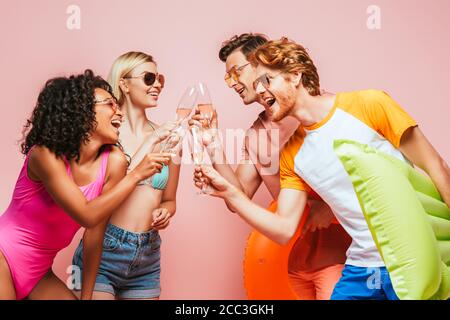 amici multiculturali eccitati in outfit estivo e occhiali da sole che clinking champagne occhiali su rosa Foto Stock