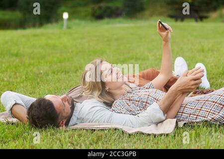 Vista laterale ritratto di una coppia spensierata che prende selfie mentre si sdraia su erba verde nel parco, copia spazio Foto Stock