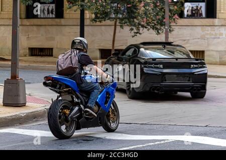 Un maschio caucasico dai capelli lunghi che indossa un bakcpack, una t-shirt, guanti, jeans e un casco sta guidando una moto sportiva in una posizione urbana. Caratteristiche dell'immagine Foto Stock