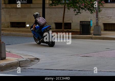 Un maschio caucasico dai capelli lunghi che indossa un bakcpack, una t-shirt, guanti, jeans e un casco sta guidando una moto sportiva in una posizione urbana. Caratteristiche dell'immagine Foto Stock