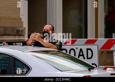 Frederick, MD, USA 08/14/2020: Un giovane poliziotto maschio caucasico indossa una maschera mentre è in servizio. È in piedi presso la sua macchina di polizia di fronte o Foto Stock