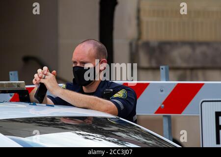 Frederick, MD, USA 08/14/2020: Un giovane poliziotto maschio caucasico indossa una maschera mentre è in servizio. È in piedi presso la sua macchina di polizia di fronte o Foto Stock