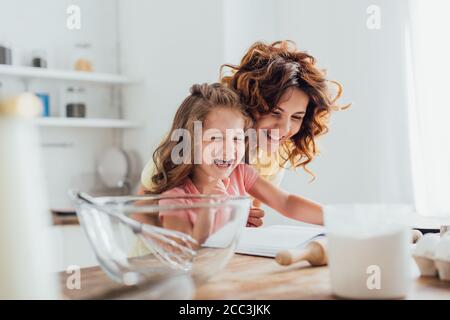 fuoco selettivo della madre e della figlia che ridono mentre leggendo il libro di cucina vicino agli ingredienti e agli utensili da cucina Foto Stock