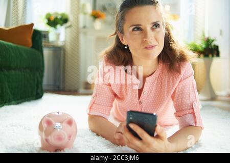 pusiva giovane casalinga in blusa e pantaloni bianchi con piggy banca alla ricerca di servizio di pulizia moquette su uno smartphone mentre posa su tappeto bianco i Foto Stock