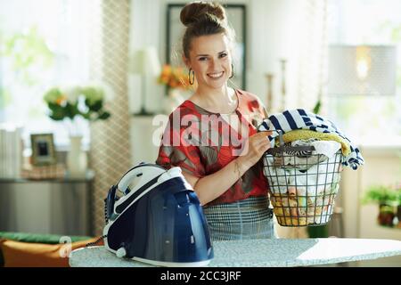 Ritratto di donna moderna felice in blusa rossa e gonna a matita grigia con cesto di vestiti lavati e ferro generatore di vapore a casa moderna in giornata di sole. Foto Stock