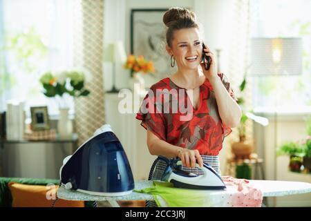 casalinga moderna sorridente di mezza età in blusa rossa e grigia stiratura con gonna a matita e generatore di vapore sull'asse da stiro utilizzo di uno smartphone in Foto Stock