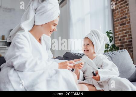 fuoco selettivo di giovane donna che fa manicure al bambino mentre sedersi insieme in accappatoi bianchi e asciugamani sulla testa Foto Stock