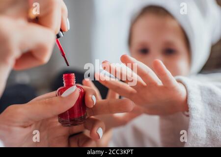 vista ritagliata della donna che applica smalto rosso sulle unghie di figlia, fuoco selettivo Foto Stock