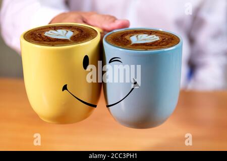 Primo piano uomo d'affari mano tenere tazze di caffè con fondo viso smle. L'uomo a mano regge tazze di caffè gialle e blu con forma a cuore cremoso e sorridente indietro Foto Stock