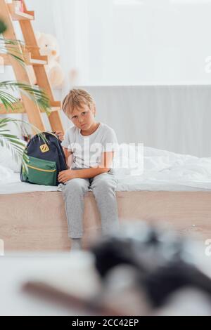 il pf di messa a fuoco selettiva sconvolse il ragazzo in pigiama guardando la fotocamera mentre si siede a letto vicino allo zaino della scuola Foto Stock