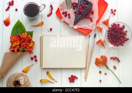 Apri un taccuino vuoto con una tazza di caffè, una fetta di torta rossa corrente, pennelli e bouquet di fiori su sfondo di legno bianco, spazio per le copie. Vista dall'alto. Foto Stock