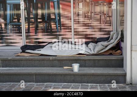 Sacco a pelo di una persona senza casa si stese sulle scale di fronte a una moderna lounge o ristorante elegante. C'è una piccola tazza di metallo vicino al berth. Foto Stock