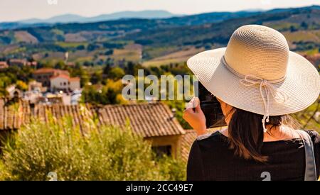 Splendida vista ravvicinata di una donna, indossando un grande cofano solare e scattando foto con il suo cellulare della bella campagna di San Gimignano in... Foto Stock