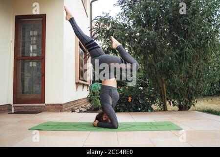 Vista laterale completa della giovane donna attiva che pratica il supporto per la testa allenati sul tappetino yoga nel patio Foto Stock