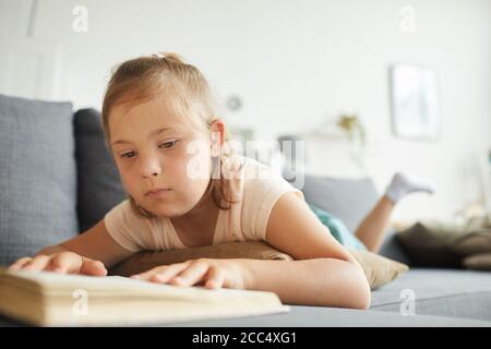 Bambina con sindrome di Down sdraiata sul divano e la lettura un libro nel soggiorno Foto Stock