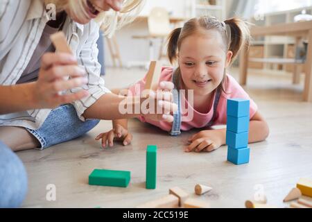 Bambina con la sindrome di Down sdraiata sul pavimento e. guardando come sua madre costruisce una torre da blocchi colorati a casa Foto Stock