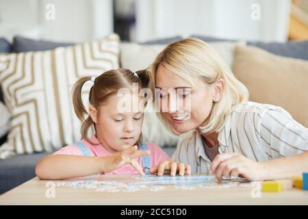 Bambina con sindrome di Down raccogliere puzzle al tavolo con l'aiuto di sua madre sono in camera a casa Foto Stock