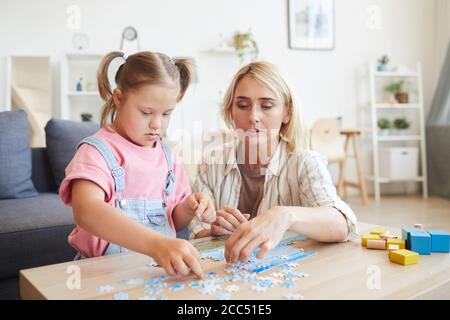 Giovane madre che aiuta sua figlia con la sindrome di Down raccolta puzzle al tavolo in camera Foto Stock