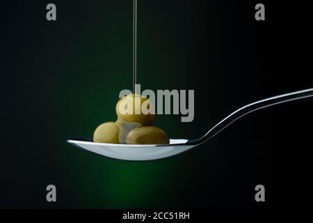 olive verdi su un cucchiaio su fondo scuro, flusso di olio d'oliva Foto Stock