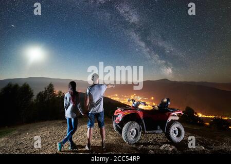 Vista posteriore di coppie felici uomini e donne con moto quad atv sulla cima della montagna. Guy che punta al bel cielo notturno pieno di stelle, Via Lattea, luna piena, luminosa città sullo sfondo Foto Stock