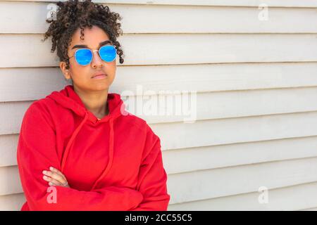 Ragazza adolescente cool teen misto razza biracial African American femmina giovane donna che indossa occhiali da sole blu e una felpa con cappuccio rossa vacanza al sole d'estate Foto Stock