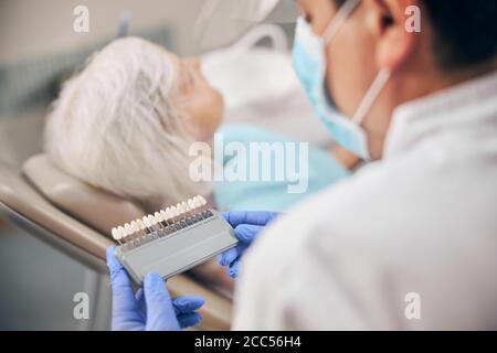 Dentista maschile che tiene in mano la tavolozza dei denti di colore per sbiancare in clinica odontoiatrica Foto Stock