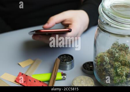Uomo che tiene il telefono accanto a cannabis e accessori per fumare. Acquisto di marijuana e erbacce online. Concetto aziendale Foto Stock