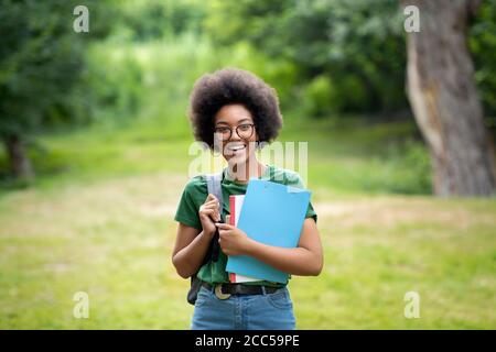 Allievo allegro dell'università femminile nera con lo zaino e i libri di lavoro che posano All'aperto Foto Stock