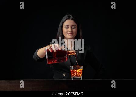 Bella barista ragazza versa una bevanda alcolica in un bicchiere con pezzi di ghiaccio. Bella brunetta in nero preparare cocktail. Spazio di copia. Foto Stock