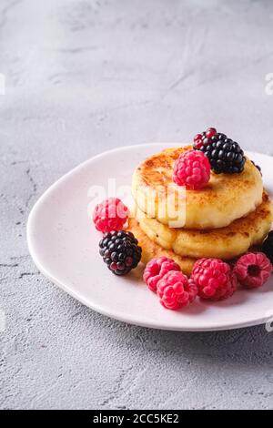 Frittelle di formaggio cottage, frittelle di cagliata dessert con lampone e bacche di mora in piastra su fondo di pietra in cemento, vista angolare Foto Stock