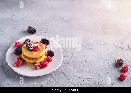 Frittelle al formaggio di cottage e zucchero in polvere, frittelle di cagliata dessert con lampone e bacche di mora in piastra su fondo di cemento di pietra Foto Stock