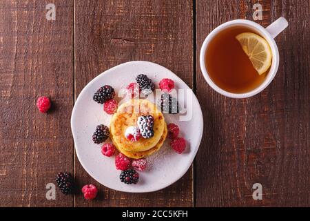 Frittelle di formaggio cottage, frittelle di cagliata dessert con lampone e frutti di bosco in piatto vicino alla tazza di tè caldo con fetta di limone Foto Stock