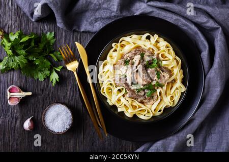 stroganoff di manzo cotto con salsa di funghi panna acida, servito con tagliatelle in una ciotola nera con forchetta dorata e coltello su un tavolo di legno scuro, fla Foto Stock