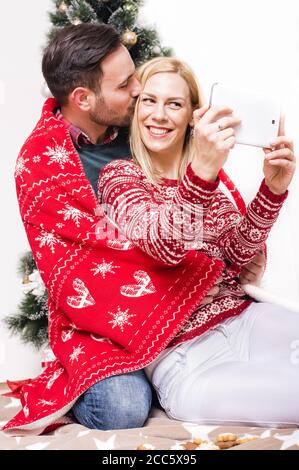 Scatto verticale di una coppia felice con una coperta rossa Prendere un selfie con un albero di Natale Foto Stock